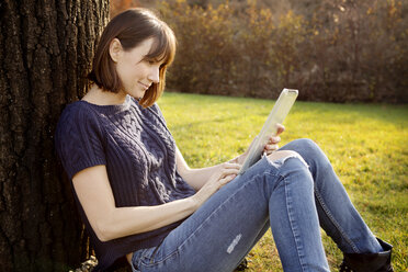 Frau benutzt Tablet-Computer, während sie im Park an einem Baumstamm sitzt - CAVF00544