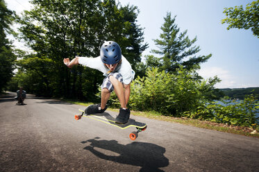 Junge fährt mit dem Skateboard über die Straße gegen die Bäume an einem sonnigen Tag - CAVF00690