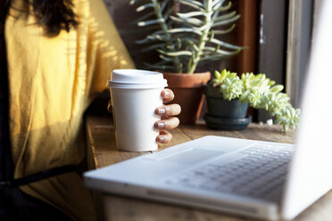 Ausgeschnittenes Bild einer Frau, die eine Kaffeetasse am Tisch mit einem Laptop zu Hause hält - CAVF00719