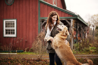 Happy woman stroking Golden Retriever while standing against barn - CAVF00741