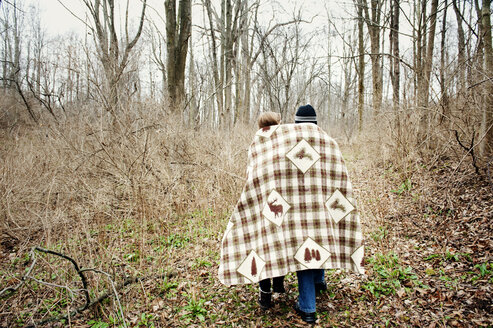 Rear view of couple wrapped in blanket while walking in forest - CAVF00748