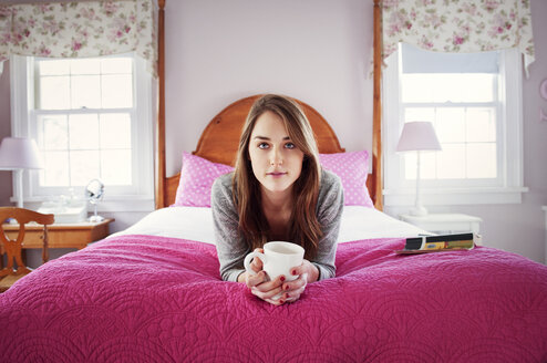 Portrait of confident woman holding coffee mug while lying on bed at home - CAVF00752