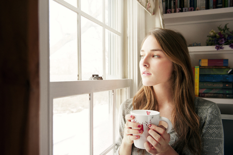 Nachdenkliche Frau, die eine Kaffeetasse in der Hand hält, während sie durch das Fenster ihres Hauses schaut, lizenzfreies Stockfoto