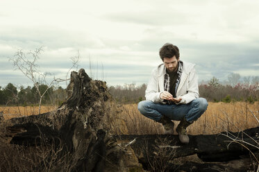 Man crouching on driftwood at field against cloudy sky - CAVF00808