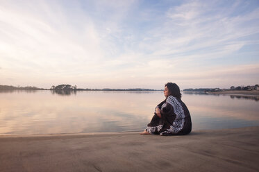 Woman wrapped in blanket while sitting on seashore - CAVF00824