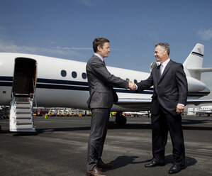 Happy businessmen doing handshake against corporate jet on runway - CAVF00856