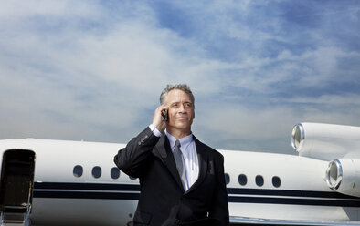 Low angle view of businessman talking on phone while standing against corporate jet on sunny day - CAVF00861