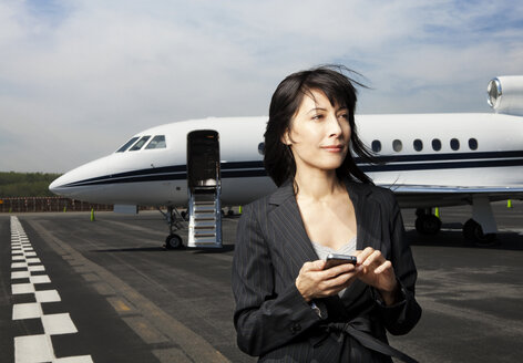 Nachdenkliche Geschäftsfrau, die ihr Telefon in der Hand hält, während sie auf der Startbahn vor einem Firmenjet steht - CAVF00862
