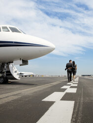 Rear view of business couple walking arms around by corporate jet against sky - CAVF00879