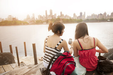 Rear view of female friends using phone while sitting at riverbank - CAVF00882