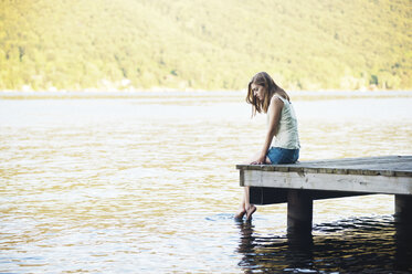Nachdenkliche Frau, die mit den Füßen das Wasser berührt, während sie auf einem Steg sitzt - CAVF00893