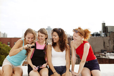 Lächelnde Frauen, die auf ihr Handy schauen, während sie auf der Terrasse eines Gebäudes gegen den klaren Himmel sitzen - CAVF00901