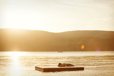 Boy lying on floating platform in lake during sunset - CAVF00992