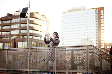 Frau fotografiert mit Smartphone auf Fußgängerbrücke vor Gebäuden - CAVF01033