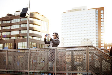 Woman photographing with smart phone on footbridge against buildings - CAVF01033