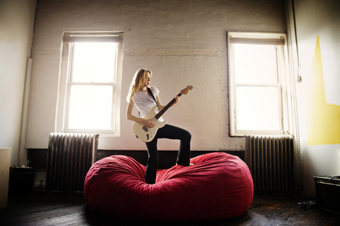 Junge Frau spielt E-Gitarre auf Sitzsack zu Hause, lizenzfreies Stockfoto