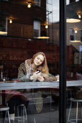 Smiling woman using smart phone in cafe seen though window - CAVF01064