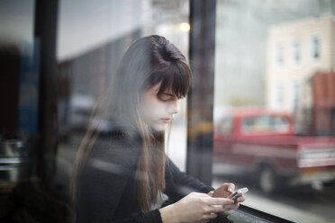 Nahaufnahme einer jungen Frau, die ein Smartphone benutzt, gesehen durch ein Fenster - CAVF01067