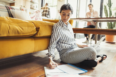 Female freelancer working at laptop, writing on paperwork in living room - CAIF04616