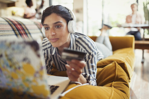 Junge Frau mit Kreditkarte und Kopfhörern beim Online-Shopping am Laptop auf dem Wohnzimmersofa - CAIF04650