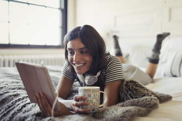 Lächelnde junge Frau, die auf dem Bett Kaffee trinkt und ein digitales Tablet benutzt - CAIF04652