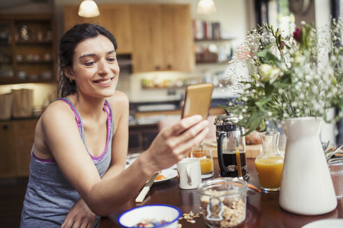 Lächelnde junge Frau, die am Frühstückstisch eine SMS mit ihrem Smartphone schreibt - CAIF04671