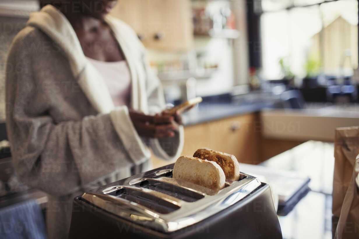 Woman texting with smart phone, toasting bread in toaster in kitchen stock  photo