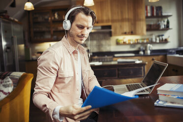 Mann mit Kopfhörern arbeitet am Laptop und liest Papierkram in der Küche - CAIF04720