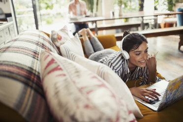 Smiling woman with headphones using laptop on sofa - CAIF04722