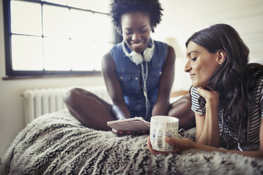Frauen mit Kopfhörern und Kaffee, die auf dem Bett ein digitales Tablet benutzen - CAIF04724