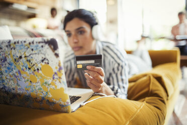 Junge Frau mit Kopfhörern und Kreditkarte beim Online-Shopping am Laptop auf dem Wohnzimmersofa - CAIF04727