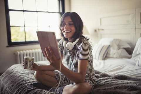 Lächelnde junge Frau mit Kopfhörern, die auf dem Bett Kaffee trinkt und ein digitales Tablet benutzt, lizenzfreies Stockfoto