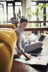 Female freelancer working at laptop, taking notes on living room floor - CAIF04733