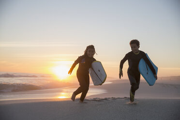 Bruder und Schwester in Neoprenanzügen laufen mit Boogie Boards auf Sommer Sonnenuntergang Strand - CAIF04782