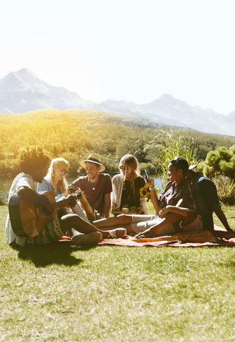 Junge Freunde hängen ab, spielen Gitarre und genießen ein Picknick im sonnigen Sommergras, lizenzfreies Stockfoto