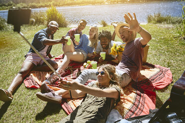 Verspielte junge Freunde machen ein Selfie mit einem Selfie-Stick und genießen ein Picknick am sonnigen Flussufer - CAIF04826