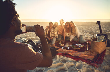 Junger Mann mit Fotohandy, der seine Freunde beim Picknick am sonnigen Sommerstrand fotografiert - CAIF04831