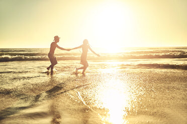Young couple holding hands, walking in sunny summer sunset ocean beach surf - CAIF04837