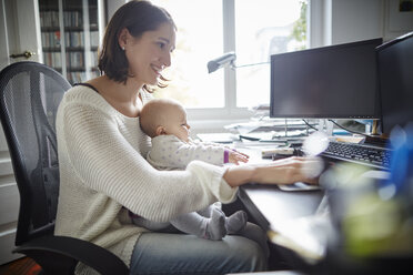 Lächelnde Mutter mit ihrer kleinen Tochter, die am Schreibtisch im Büro arbeitet - CAIF04934