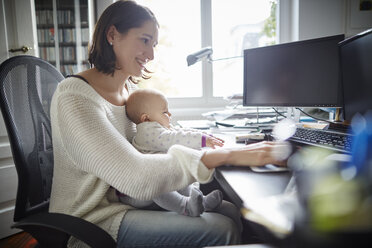 Smiling mother holding baby daughter working at desk in home office - CAIF04934
