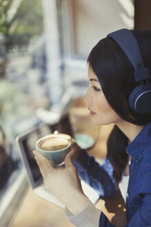 Nachdenkliche junge Frau trinkt Kaffee und hört Musik mit Kopfhörern am Fenster eines Cafés - CAIF04984