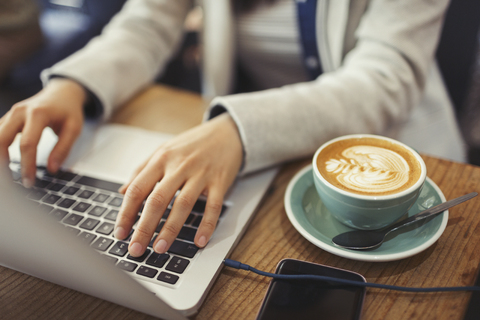 Hände einer jungen Frau, die einen Laptop benutzt und Cappuccino in einem Café trinkt, lizenzfreies Stockfoto