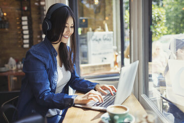 Junge Frau mit Kopfhörern und Laptop am Fenster eines sonnigen Cafés - CAIF05009