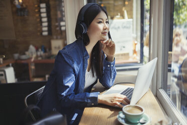 Nachdenkliche junge Frau, die mit Kopfhörern am Laptop Musik hört und in einem Café am Fenster Kaffee trinkt - CAIF05012