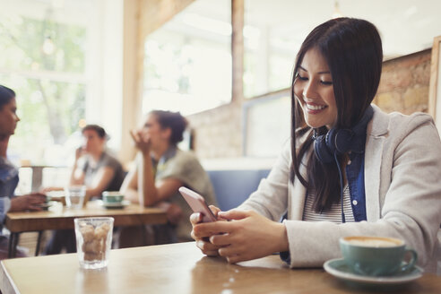 Lächelnde junge Frau mit Kopfhörern, die mit ihrem Handy SMS schreibt und Kaffee am Kaffeetisch trinkt - CAIF05021