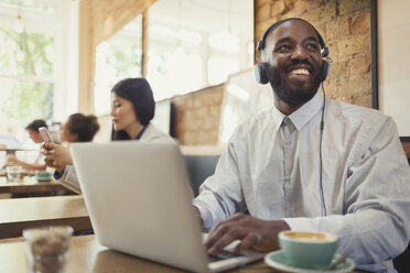 Lächelnder junger Mann mit Kopfhörern, der einen Laptop benutzt und am Cafétisch Kaffee trinkt - CAIF05023