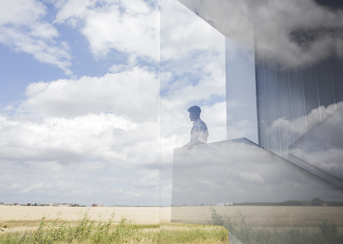 Nachdenklicher Geschäftsmann auf einem modernen Treppenabsatz, der aus dem Fenster auf den sonnigen blauen Himmel und die Wolken schaut, lizenzfreies Stockfoto