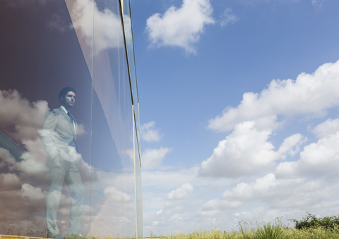 Nachdenklicher Geschäftsmann blickt aus einem modernen Bürofenster auf einen sonnigen blauen Himmel und Wolken, lizenzfreies Stockfoto