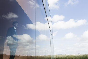 Nachdenklicher Geschäftsmann schaut aus einem modernen Bürofenster auf blauen Himmel und Wolken - CAIF05034