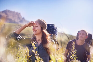 Smiling couple with backpacks hiking in sunny field - CAIF05079